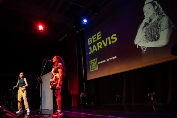 A side-profile of a musician, Bee Jarvis, on-stage. Bee is singing into a microphone and has a look of concentration on her face, with her eyes closed. Her arms are by her side, and she wears a colourful dress.