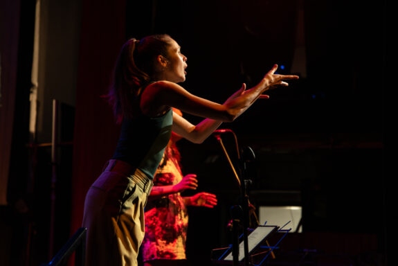 Side-profile of a BSL interpreter on-stage. Her arms are outstretched in front of her, nearly at a 90 degree angle. Behind her is an artist on stage.