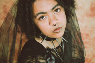 A Black-Asian person in their early twenties with dark brown dreadlocks, a septum piercing and a black chocker with spikes. They are wearing a black veil, and are looking up at the camera.