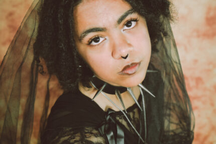 A Black-Asian person in their early twenties with dark brown dreadlocks, a septum piercing and a black chocker with spikes. They are wearing a black veil, and are looking up at the camera.