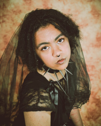 A Black-Asian person in their early twenties with dark brown dreadlocks, a septum piercing and a black chocker with spikes. They are wearing a black veil, and are looking up at the camera.