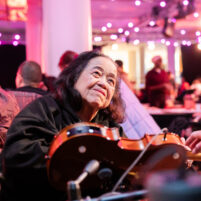 A Disabled adult playing an adapted violin instrument that is mounted to a table top. They are smiling warmly.