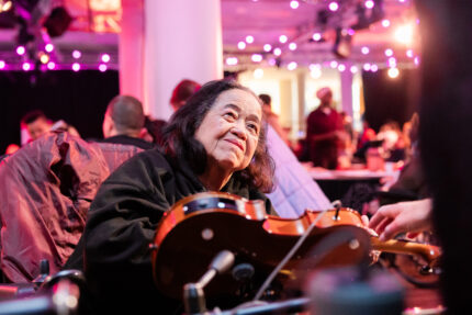 A Disabled adult playing an adapted violin instrument that is mounted to a table top. They are smiling warmly.