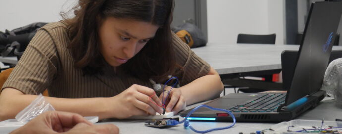 A hackathon participant working with electrical components and a laptop on a desk. They have a concentrated look on their face.
