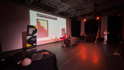A shot of a large event room. In the center is a person sat down at a table, presenting with a projection screen behind them. There is also a table with accessible instruments laid out, a table with people sat down and a BSL interpreter standing and signing.