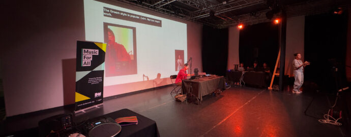 A shot of a large event room. In the center is a person sat down at a table, presenting with a projection screen behind them. There is also a table with accessible instruments laid out, a table with people sat down and a BSL interpreter standing and signing.
