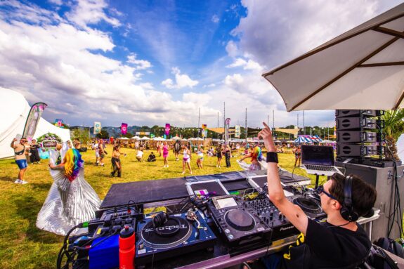 A photo taken from behind a DJ who is a wheelchair user. His left arm is raised in the air, as he looks straight ahead at a crowd of dancers who are gathered on a large area of grass.