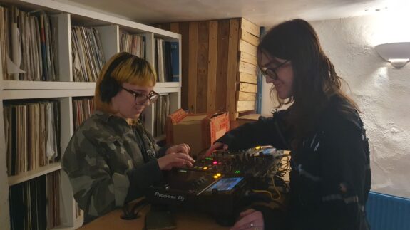 Lilian guiding a workshop participant, showing them how to operate DJ decks. They are in a room with a low ceiling, behind them is a large collection of records.