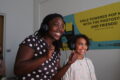 An adult and child standing side-by-side, smiling. They both wear headphones and are in a room with white walls.