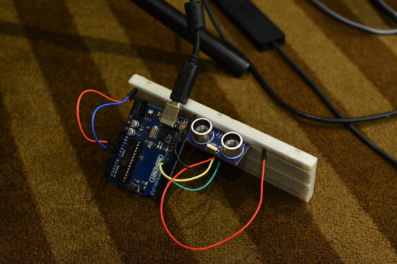 An electronic device made up of an arduino circuit board, and red, blue, green, yellow and black wires. It lies on a stripey brown carpet.