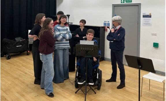 A group of young students and learner are stood around Ale, one of the members of The Clarion Trio. They are smiling, and are looking at the equipment.