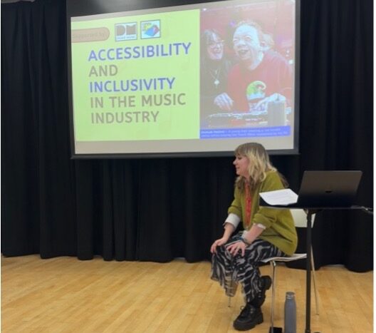 Pea, a young person, sat on a chair in front of a screen which reads 'accessibility and inclusivity in the music industry'.