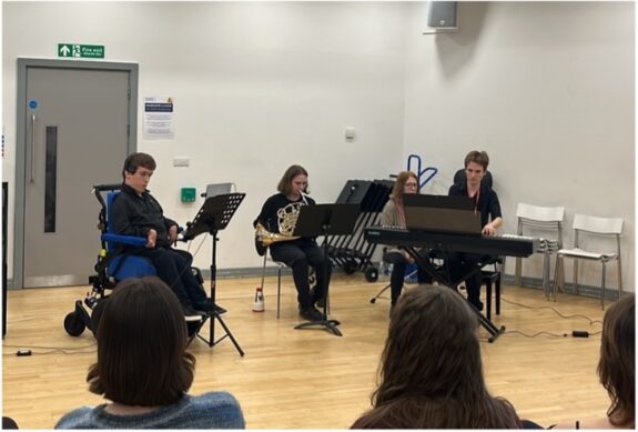 The Clarion Trio, made up of three young Disabled people, sat performing in a classroom setting. One person plays the keys, another the French horn, whilst the other plays an accessible instrument.