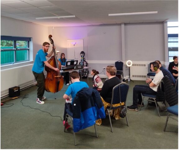 A group of young people sat in a classroom on chairs, using accessible instruments. A teacher is at the front, playing a double bass or cello. 