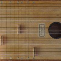 Picture of wooden musical instrument taken from above. The instrument is rectangular and has 5 sets of 6 strings, each with a bridge located at different points up the length of the strings. There is a sound hole and to the left of the sound hole there is a label which says “Pentachord”. Each group of strings is labelled with a different coloured dot sticker, and there are lines going across like frets labelled with note names at the top and bottom.