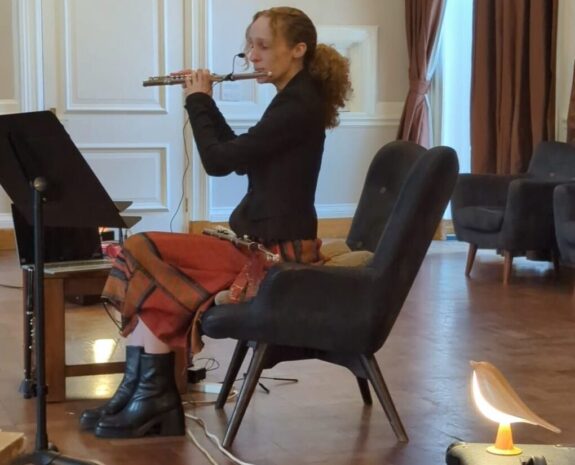 Amélie, a young person, sat down on an armchair performing the flute. The room is large and comfortable-looking, with lots of natural and warm lighting.