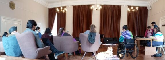 A photo taken from behind the audience at Amelie's concert. Members are sat down in large armchairs, with one person in a wheelchair. Large brown curtains cover the windows, and 3 small chandeliers lights the room.