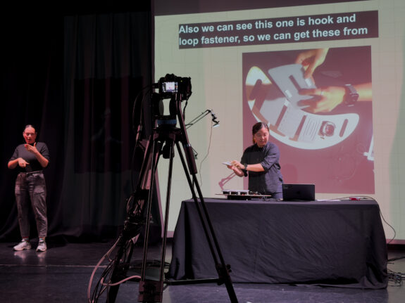 A camera mounted on a tripod, pointing towards a person who is presenting an accessible instrument on stage. Behind them is a projector screen, which shows a close up of an instrument. To their right is a BSL interpreter.