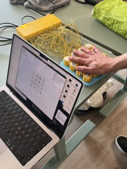 A hand pressing on a handmade electronic palm-sized instrument made of circular yellow conductors in rows and lots of yellow cables coming out of a box and connected to a laptop controlling it