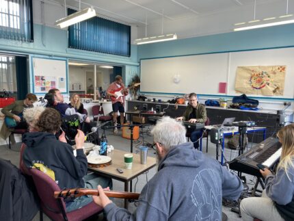 A group of Disabled and Non-Disabled people playing instruments in a group. They are in a classroom which is brightly lit.