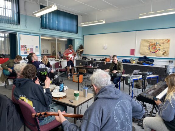 A group of Disabled and Non-Disabled people playing instruments in a group. They are in a classroom which is brightly lit.