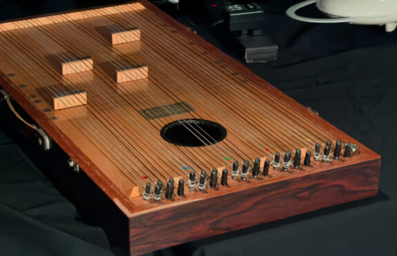 A rectangular wooden instrument with rows of around 30 strings running lengthways across it, with black pegs at one end.