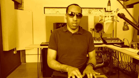 A man wearing sunglasses is facing the camera in a music studio setting. He is playing an instrument which is out of view of the camera.
