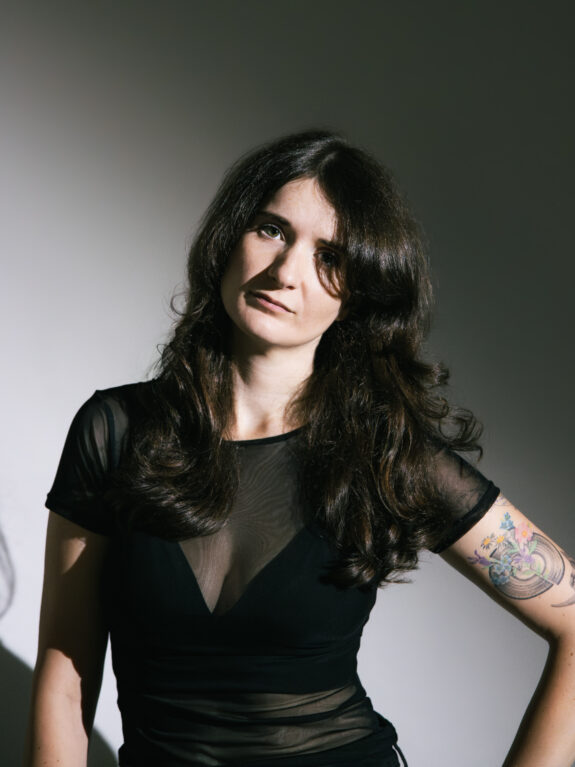 A white woman with dark hair down to their chest looks at the camera, They wear all black and have a tattoo of a vinyl record and flowers.