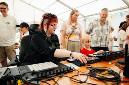 A Disabled musician leading a music workshop with young children. They are sat at a table inside a bright marquee, with musical equipment laid out infront.