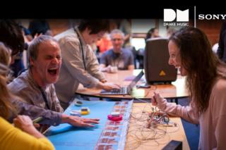 5 people sat and stood around a table which has various electronic equipment and other materials laid across it. They are laughing, and the mood is light. There are white logos for Drake Music and Sony in the top right corner.