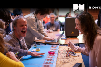 5 people sat and stood around a table which has various electronic equipment and other materials laid across it. They are laughing, and the mood is light. There are white logos for Drake Music and Sony in the top right corner.