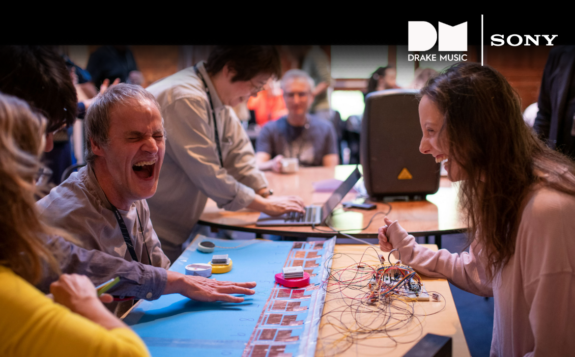 5 people sat and stood around a table which has various electronic equipment and other materials laid across it. They are laughing, and the mood is light. There are white logos for Drake Music and Sony in the top right corner.