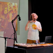 Billy Payne, a white man with long dark hair and a beard. He is presenting, with a projection screen behind him. A rectangular wooden instrument lies on the desk in front of him.