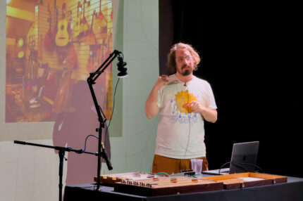 Billy Payne, a white man with long dark hair and a beard. He is presenting, with a projection screen behind him. A rectangular wooden instrument lies on the desk in front of him.