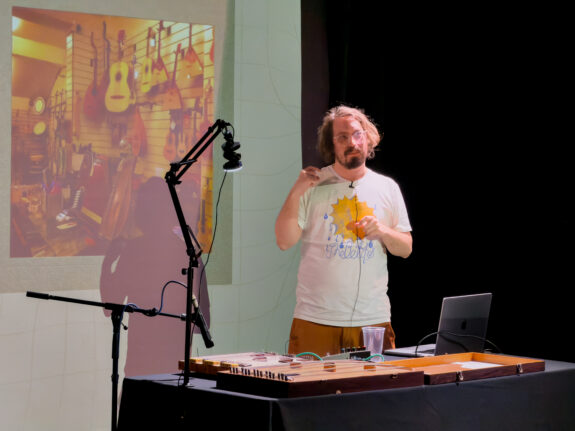 Billy Payne, a white man with long dark hair and a beard. He is presenting, with a projection screen behind him. A rectangular wooden instrument lies on the desk in front of him.