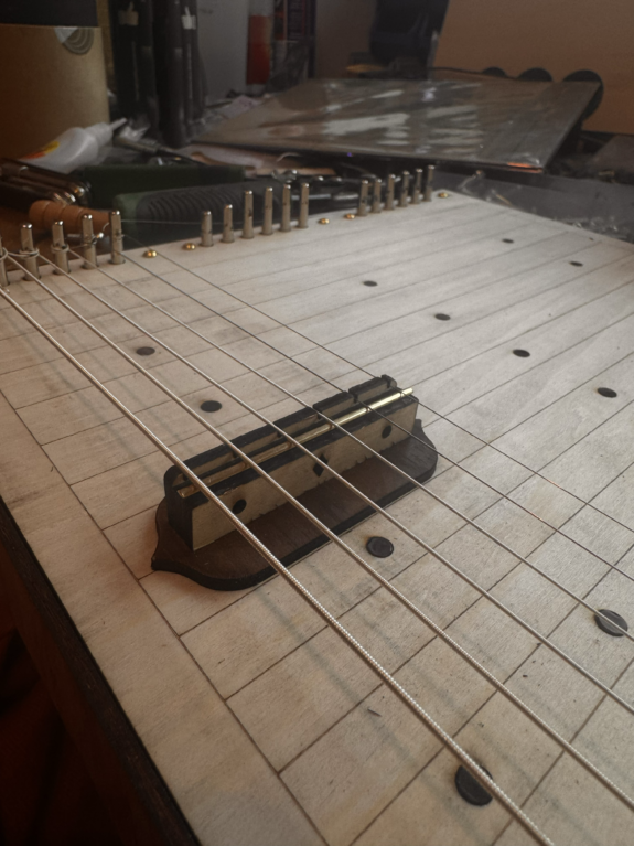 A close up images of 6 strings draw across the Pentachord. The strings are thick, and are secured using dials at either end. It is attached to pale wood.